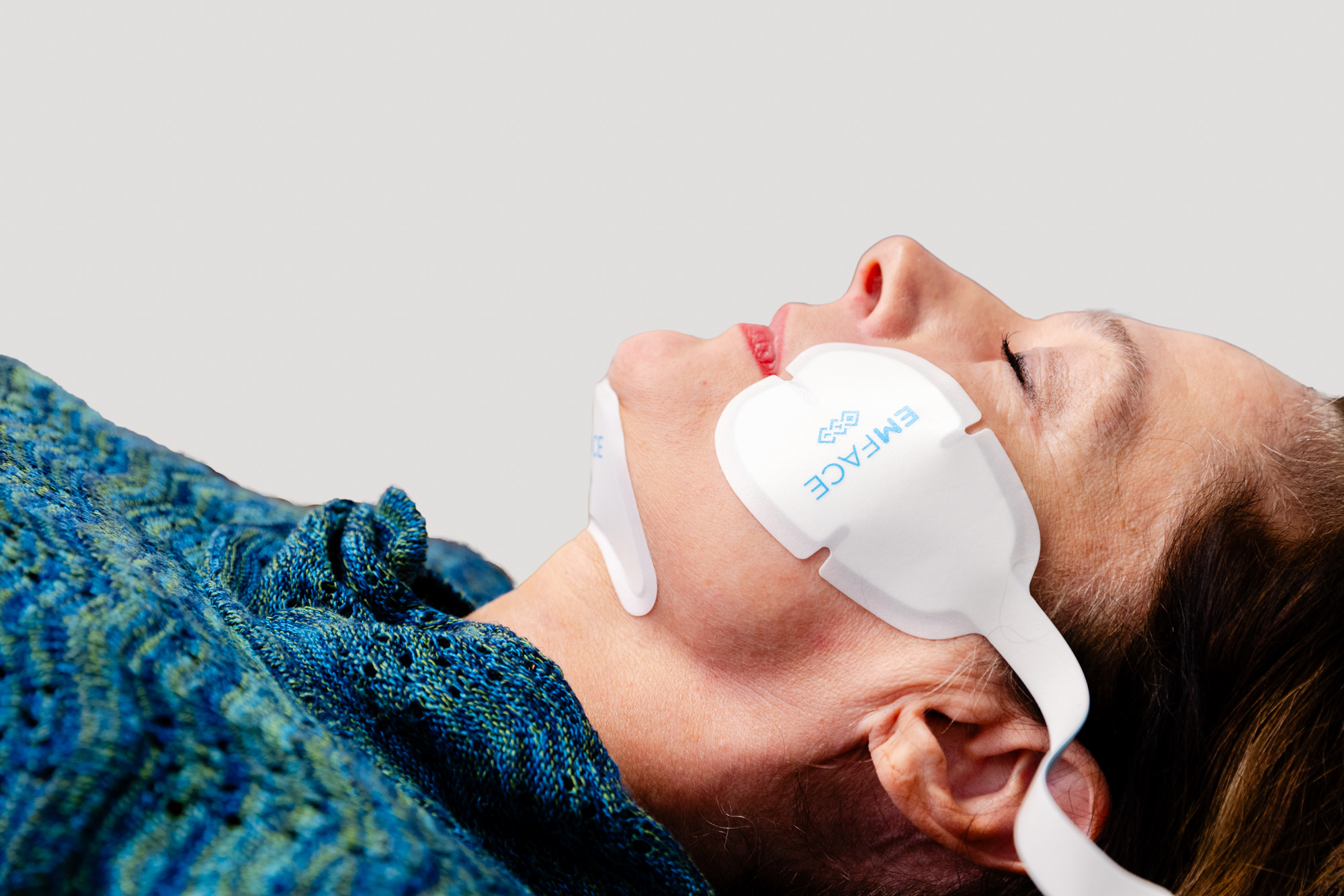 Sideview of a owmans face as she relaxes on a treatment table. She has applicators attached to her cheeks and under her chin, part of her Emface anti-aging treatment in Newburgh.